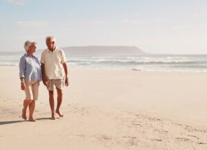 Elderly Couple Walking on a Quiet Beach Senior couple walking hand in hand along a peaceful sandy beach with gentle waves and distant hills under soft sunlight.